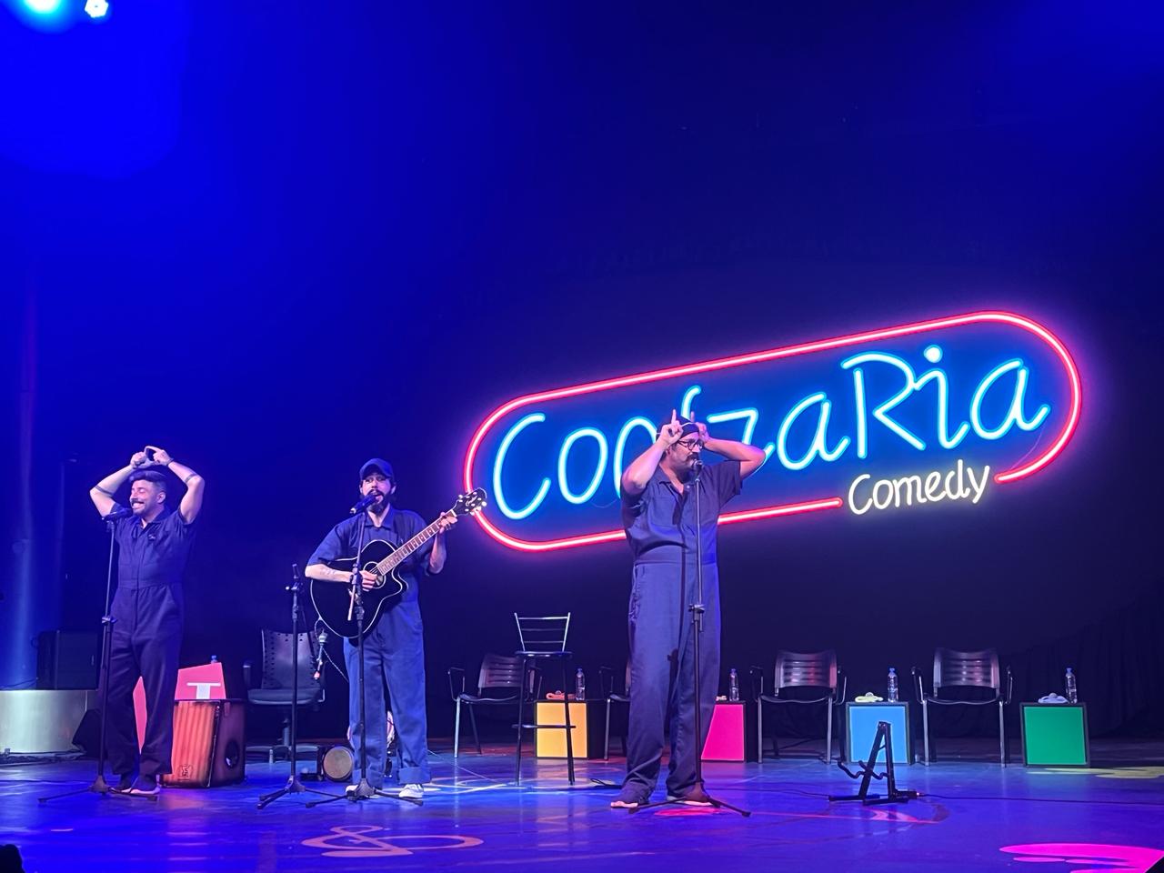 O trio no palco sob o letreiro de neon da Confraria Comedy